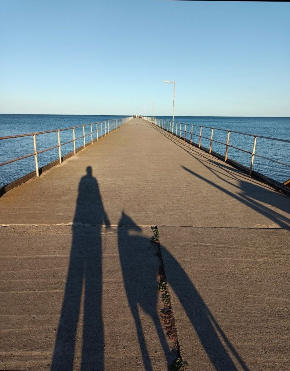 A long concrete jetty stretching out over calm sea, with the long shadows of Colleen and Winter cast across it in the low sun.